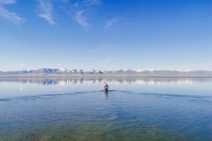 A figure wades into a lake under a clear sky