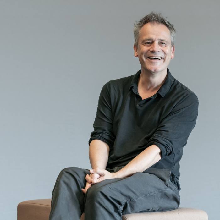 Photographic portrait of Michael Grandage sitting on a stool in a studio, laughing